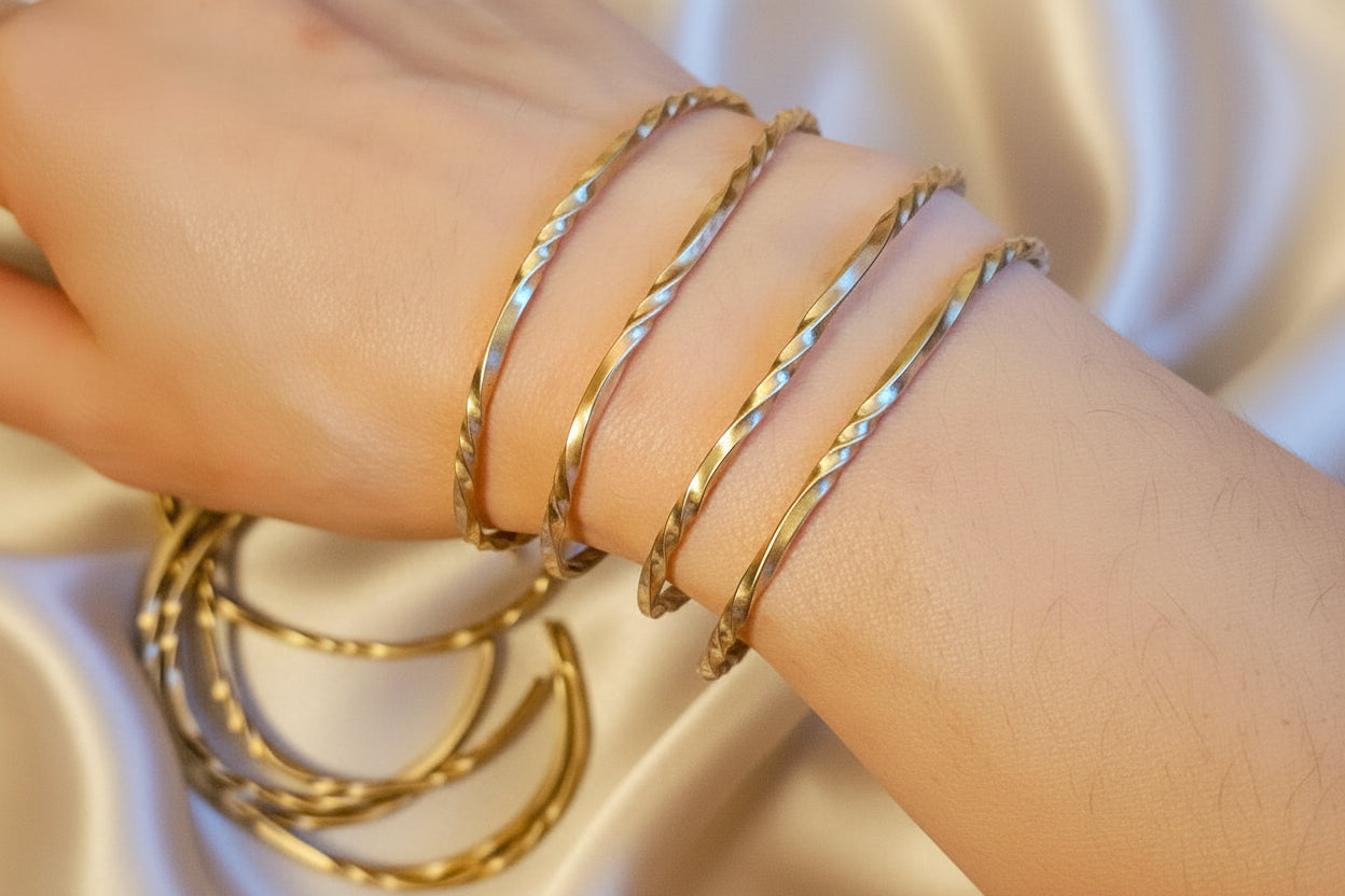 Close-up of a wrist wearing multiple ribbon cuff gold bracelets on a textured fabric background
