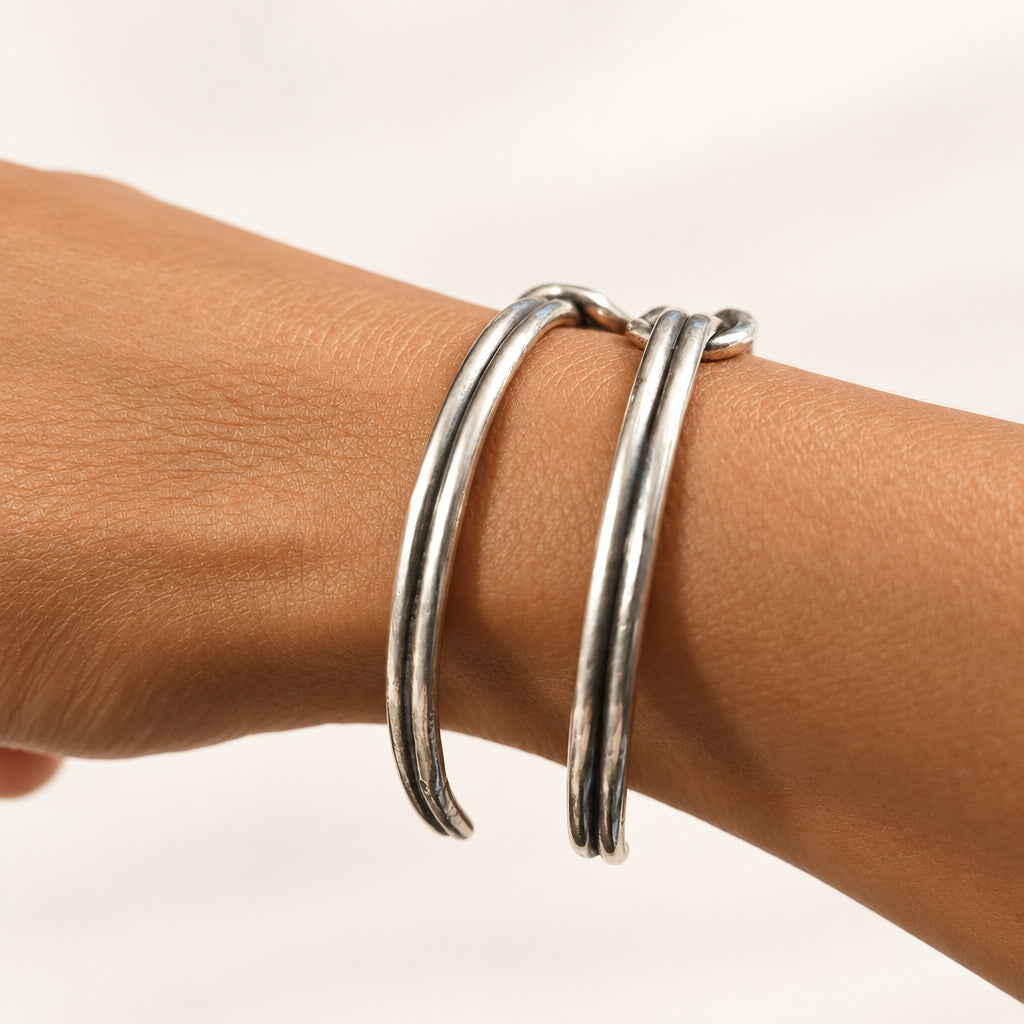 A close-up of a person's wrist wearing two polished silver bangle bracelets, with a blurred indoor background.